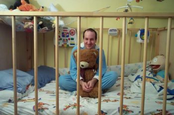 A Man Sitting Inside Baby Cot along with teddy bear